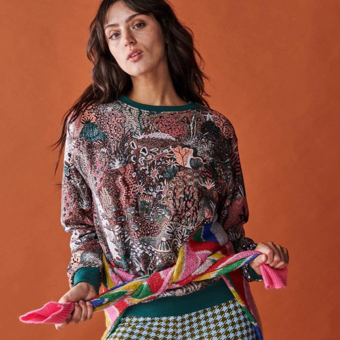 Model is tall with long brown layered hair and is looking into the camera. The models wears a Deep Sea Organic jumper which features green, maroon and pink coloured underwater images. The model is wearing blue and brown hounds tooth leggings and has tied a rainbow colour jumper around her waist.